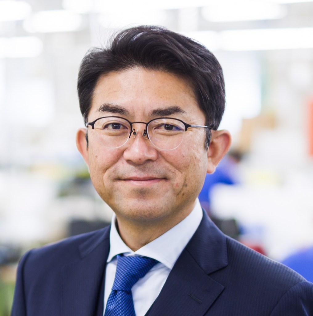 Portrait of a businessman wearing glasses and a suit, smiling directly at the camera against a blurred background.